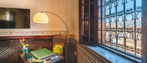 Una habitación con un escritorio de madera, libros, un florero con flores, una lámpara y vistas a través de una ventana con barrotes.