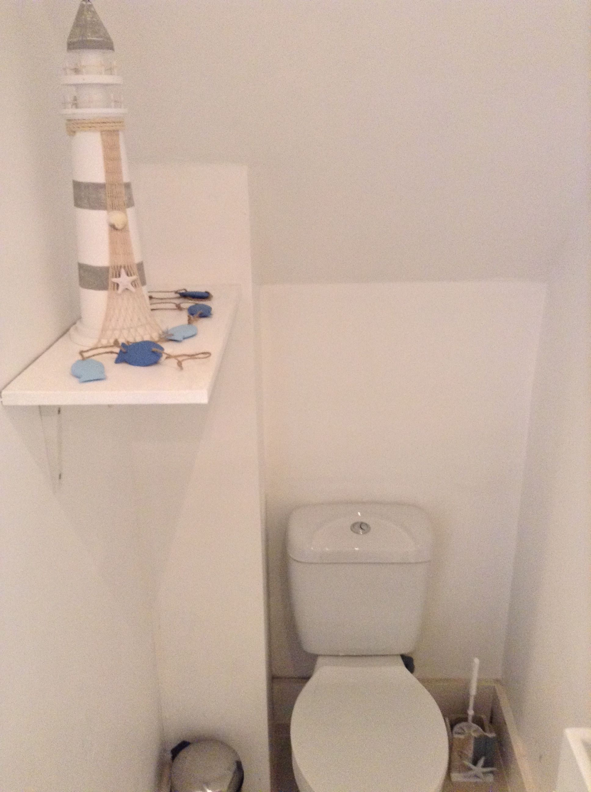 A bathroom with a toilet, a small trash bin, and a shelf with a decorative lighthouse model.