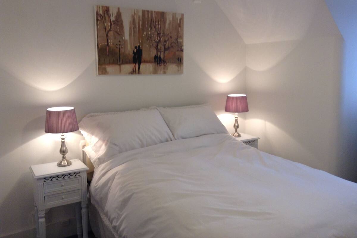 A bedroom with a white bed, two bedside lamps, and a painting on the wall.