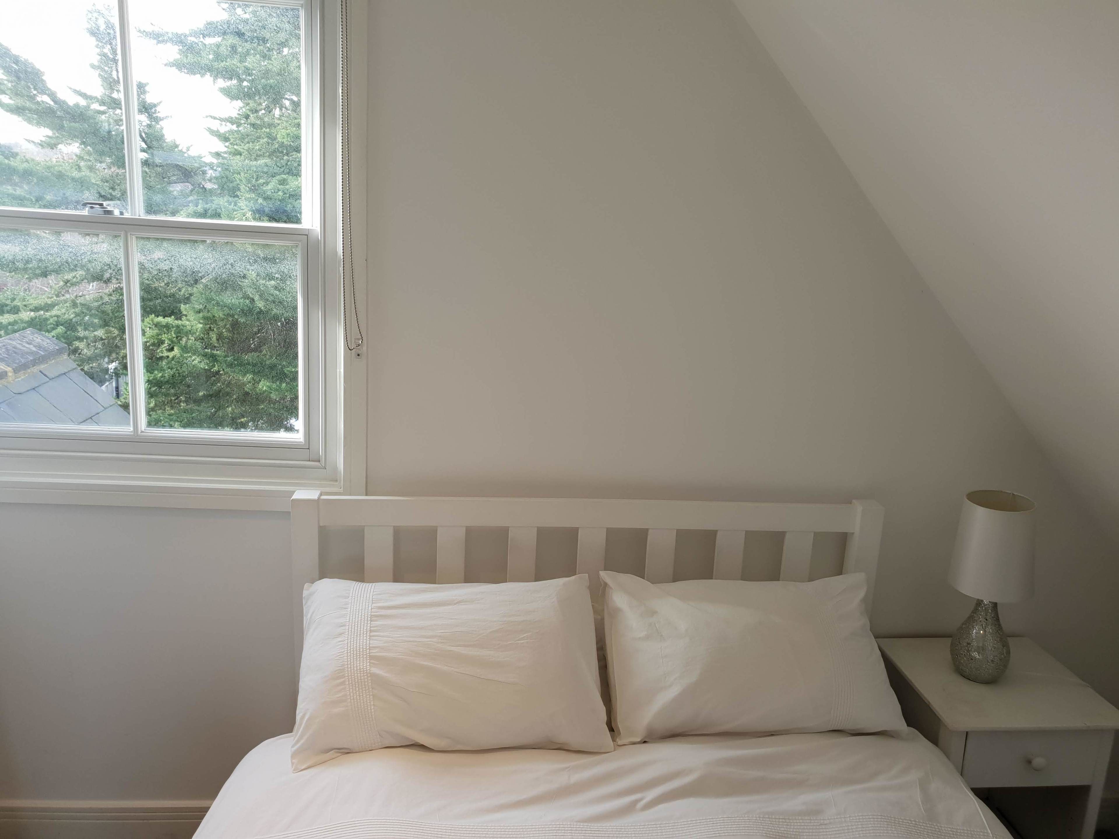 A bedroom with a white bed, a nightstand with a lamp, and a window with a view of trees.
