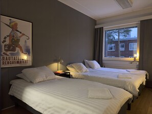 Habitación de hotel con dos camas, una mesita de noche, una lámpara, un cartel y una ventana con vistas a edificios.