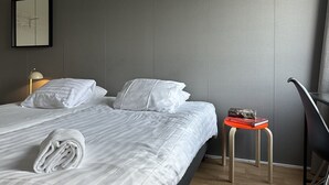 Una cama bien hecha con sábanas blancas, una toalla doblada y una mesita con un libro y una bandeja.