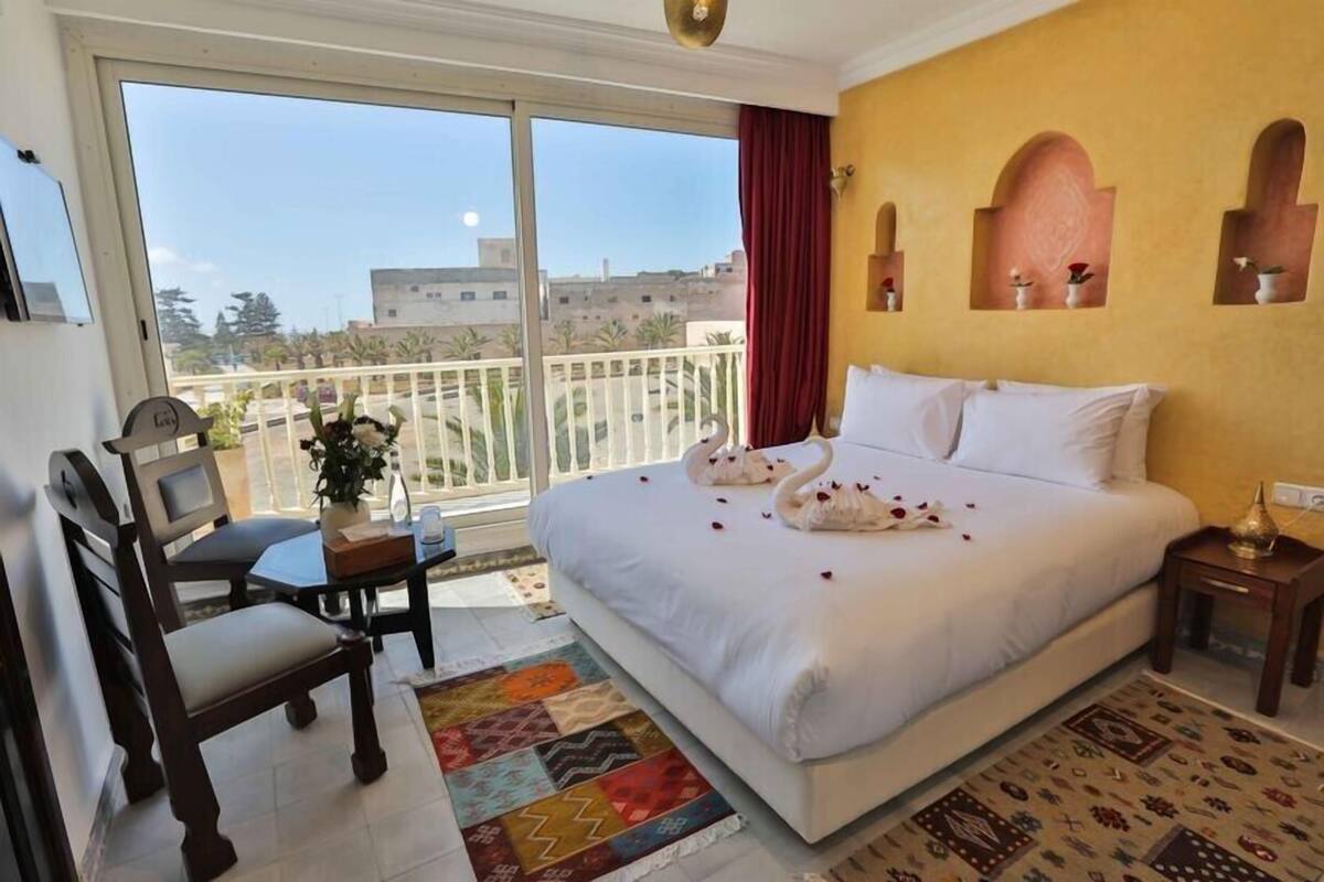 A hotel room with a large bed, a balcony with a view, a small table with a plant, and a chair.