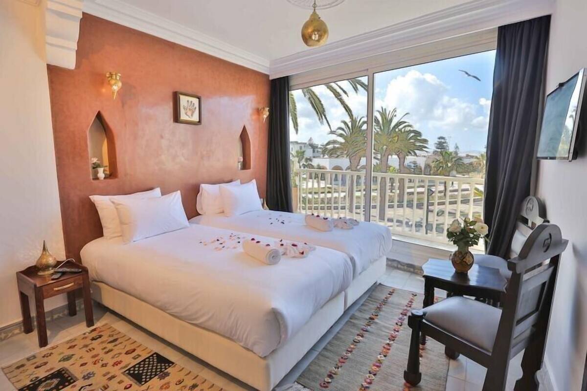 A hotel room with a large bed, a small table with a vase, and a view of palm trees and a balcony.