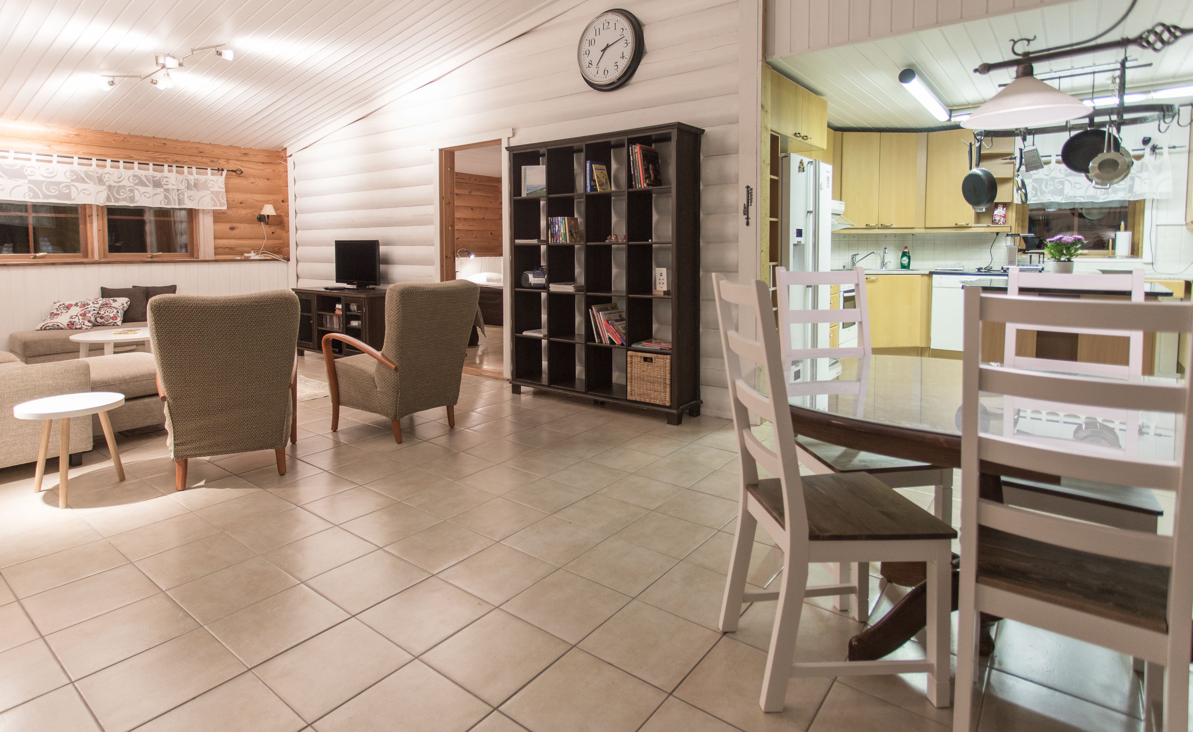 Una sala de estar con un sofá, dos sillones, una mesa de centro, una estantería, un televisor y una cocina con una mesa de comedor y sillas.