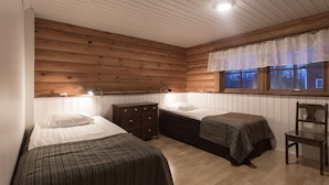 Ein schlicht eingerichtetes Schlafzimmer im Chalet-Stil mit zwei Betten, Holzwänden, einem Fenster mit Vorhängen, einem Schminktisch und einem Sessel.