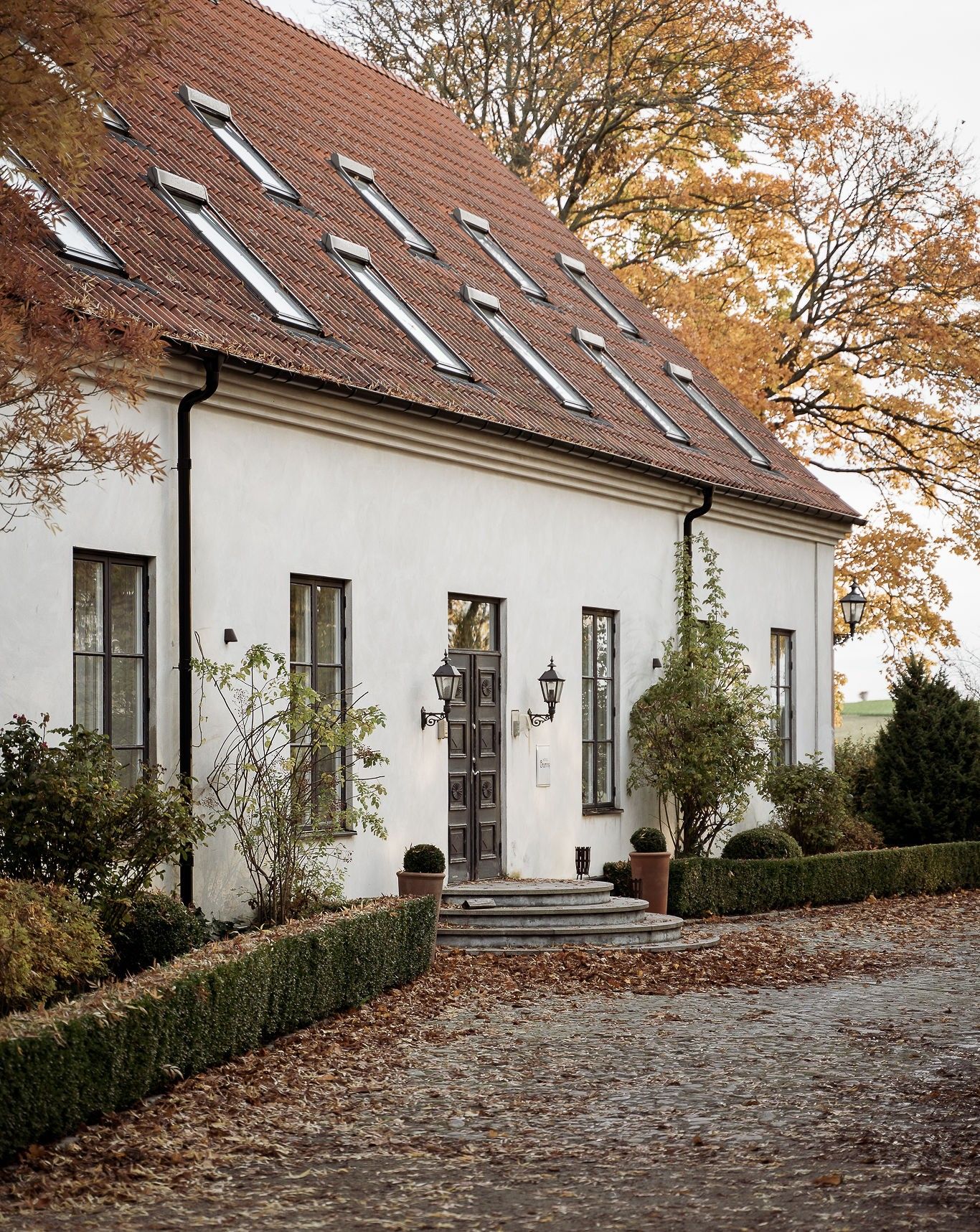 A white building with a tiled roof, a central entrance with a door, and a small garden area with a hedge and a potted plant.