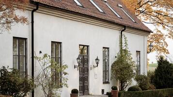 A white building with a tiled roof, a central entrance with a door, and a small garden area with a hedge and a potted plant.