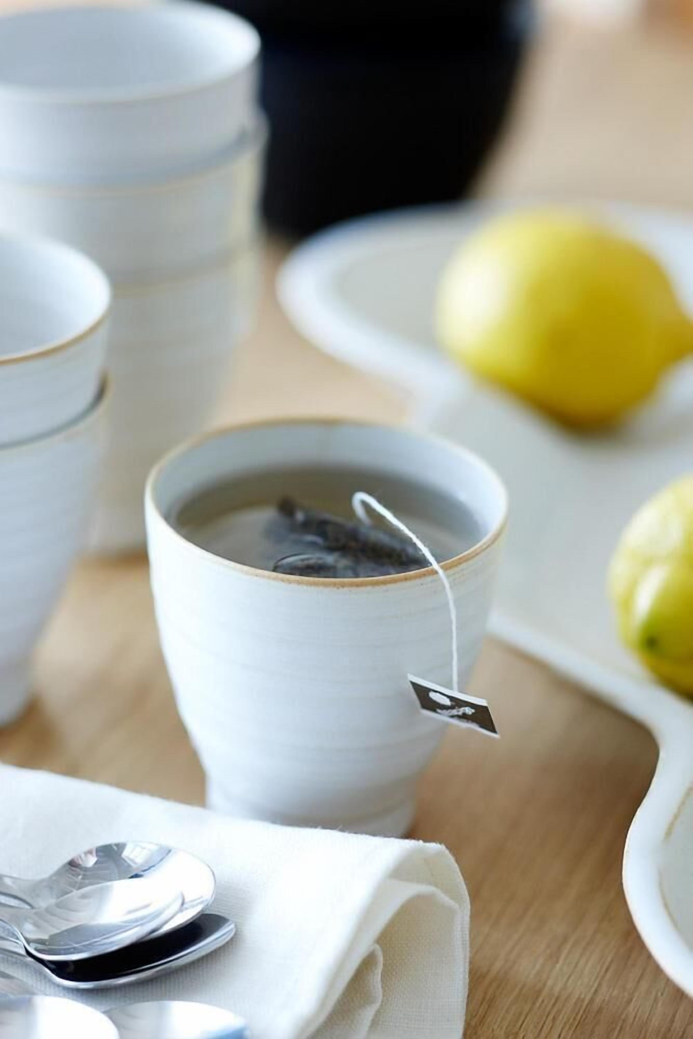 Una taza de cerámica blanca con una bolsita de té, una cuchara y un plato con limones sobre una mesa de madera.