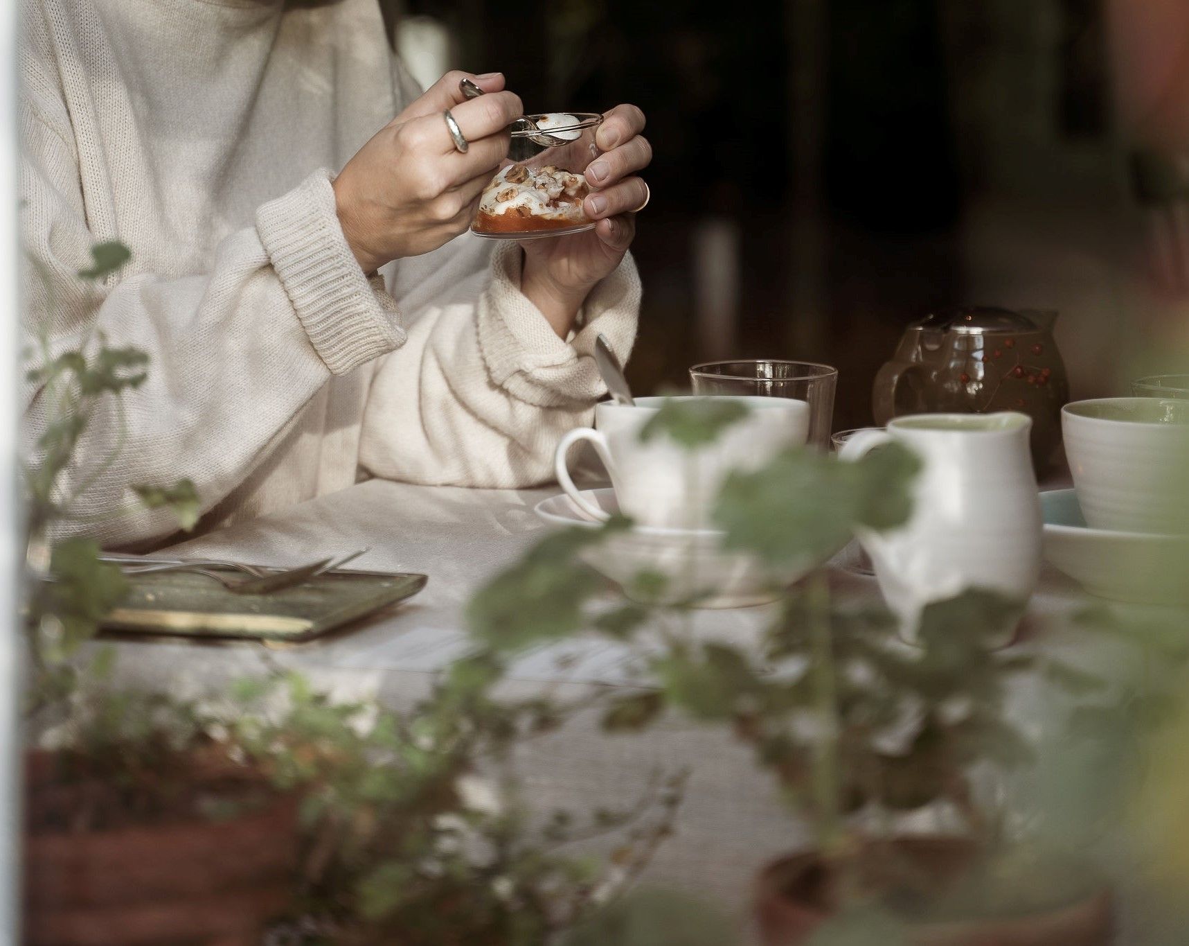 A person holding a glass with a dessert, seated at a table with various cups and a teapot.