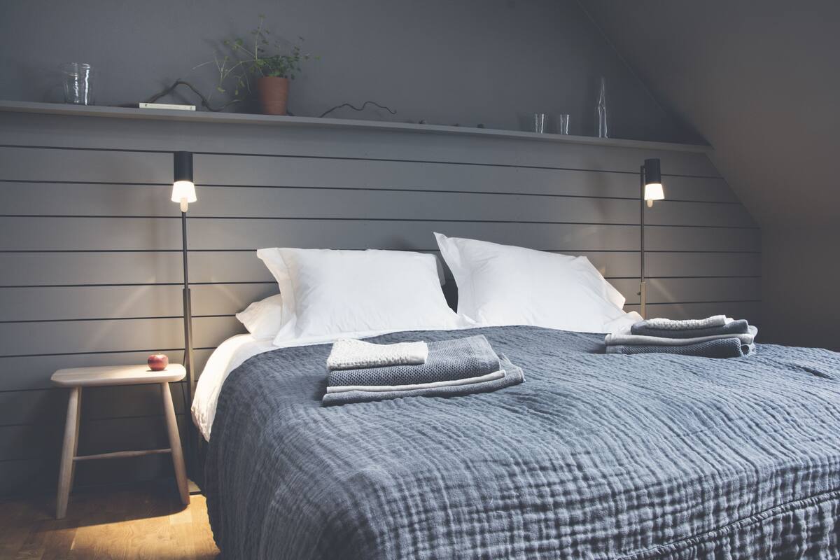 A neatly made bed with a grey checkered duvet, two white pillows, and a small stool with a red object beside it.