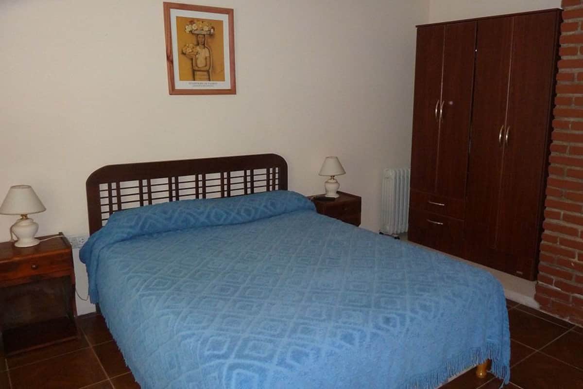 A bedroom with a bed, two bedside tables, a nightstand, a wooden wardrobe, and a brick wall.