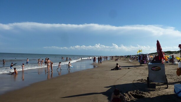 A crowded beach with people swimming and walking along the shore. - Merlión (Mar de Las Pampas)