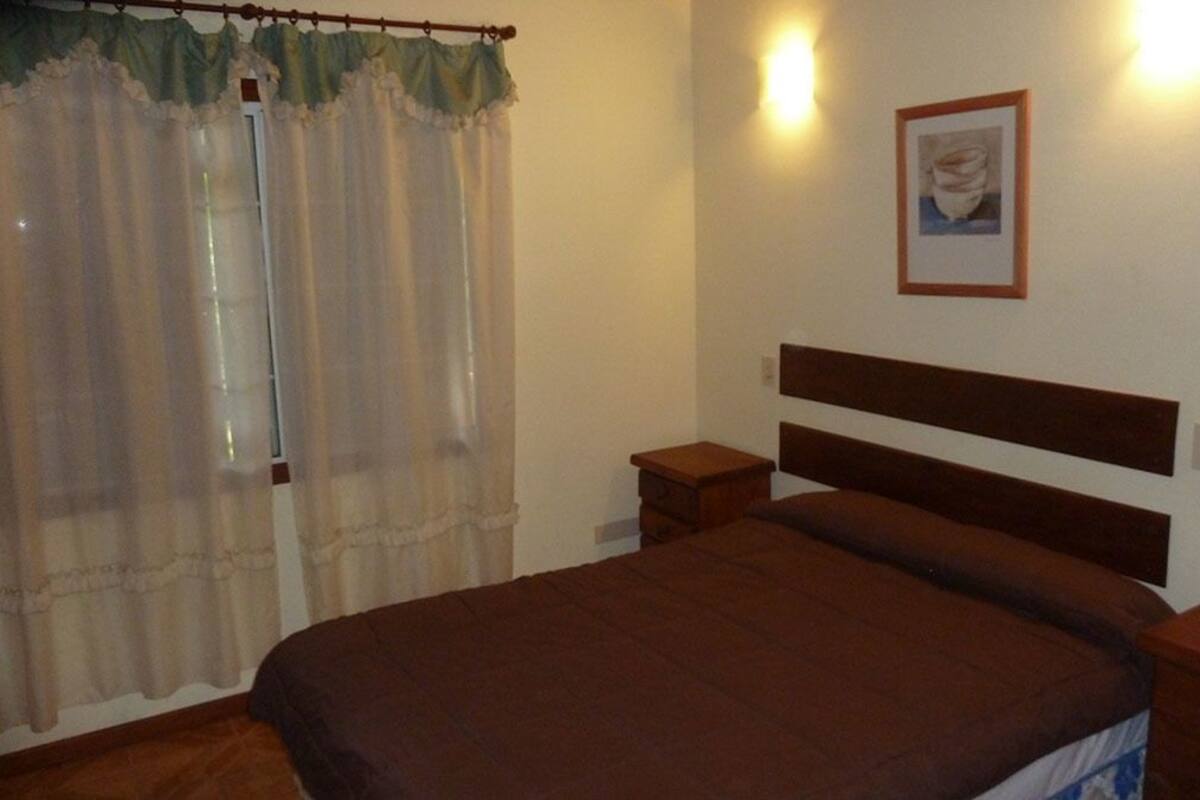 A bedroom with a single bed, a nightstand, a bedside table, a framed picture on the wall, and a window with sheer curtains.