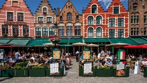 Un café al aire libre concurrido, con arquitectura europea tradicional, mesas en la terraza y un cielo azul despejado.