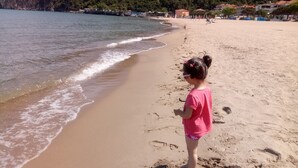 A beach with a child standing on the sand, waves gently lapping at the shore, and buildings in the background. - Kure Dag Evi (Ulus)