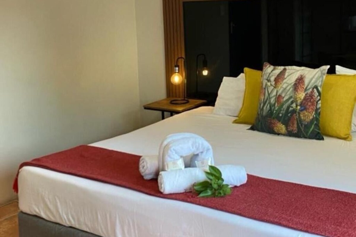 A neatly made bed with a red blanket, white pillows, and a floral pillow. There is a small table with a lamp beside the bed.