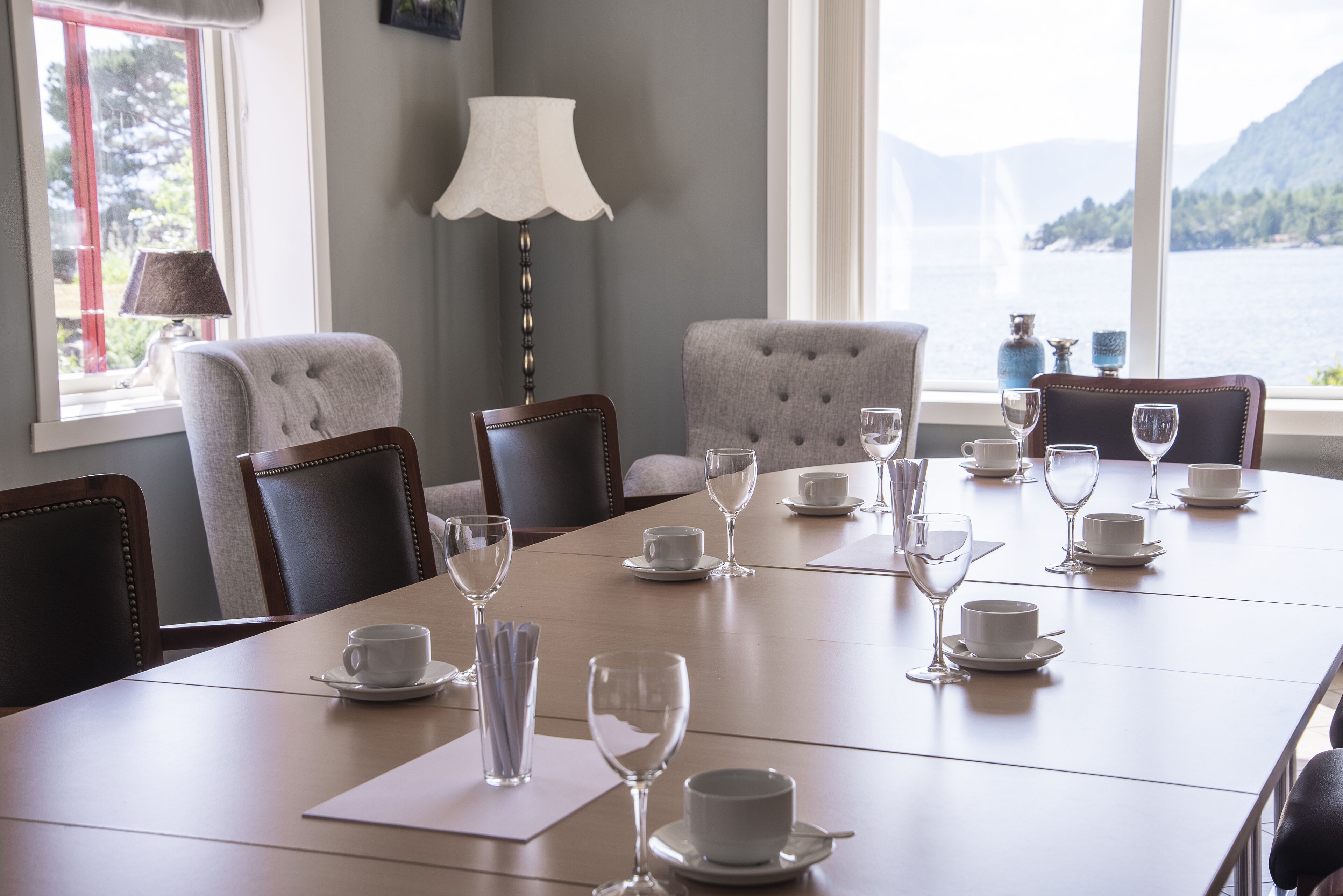 A dining table set with white cups and wine glasses, surrounded by upholstered chairs, with a view of a lake and mountains through large windows.