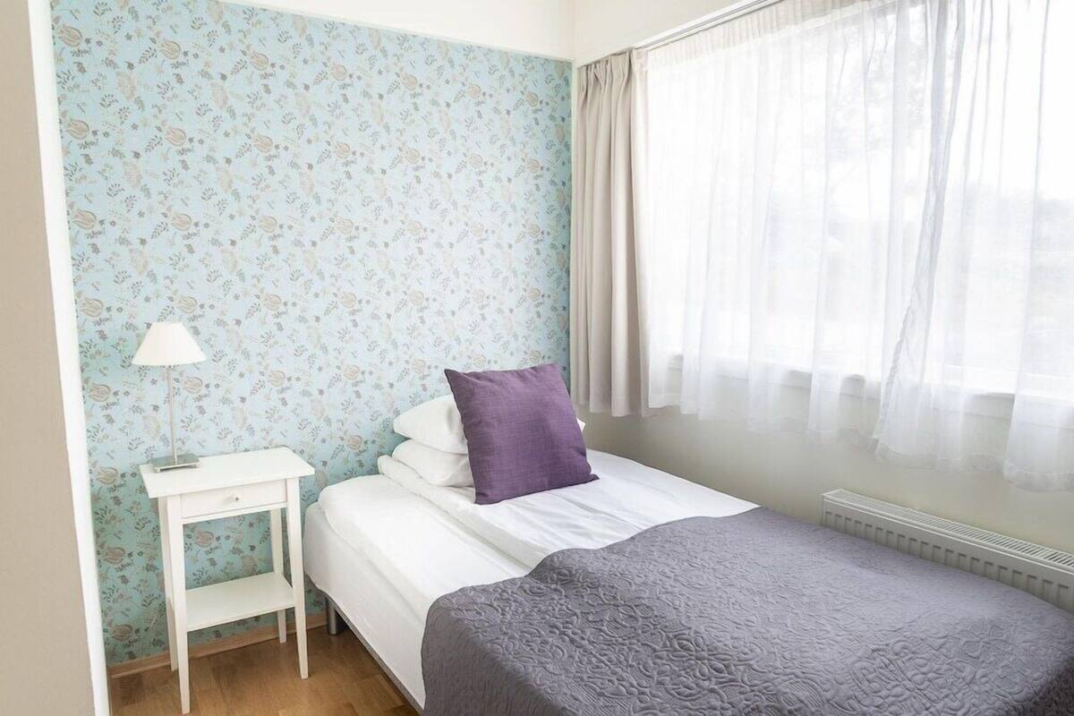 Una camera da letto con un letto, un comodino, una lampada e una finestra con tende leggere.