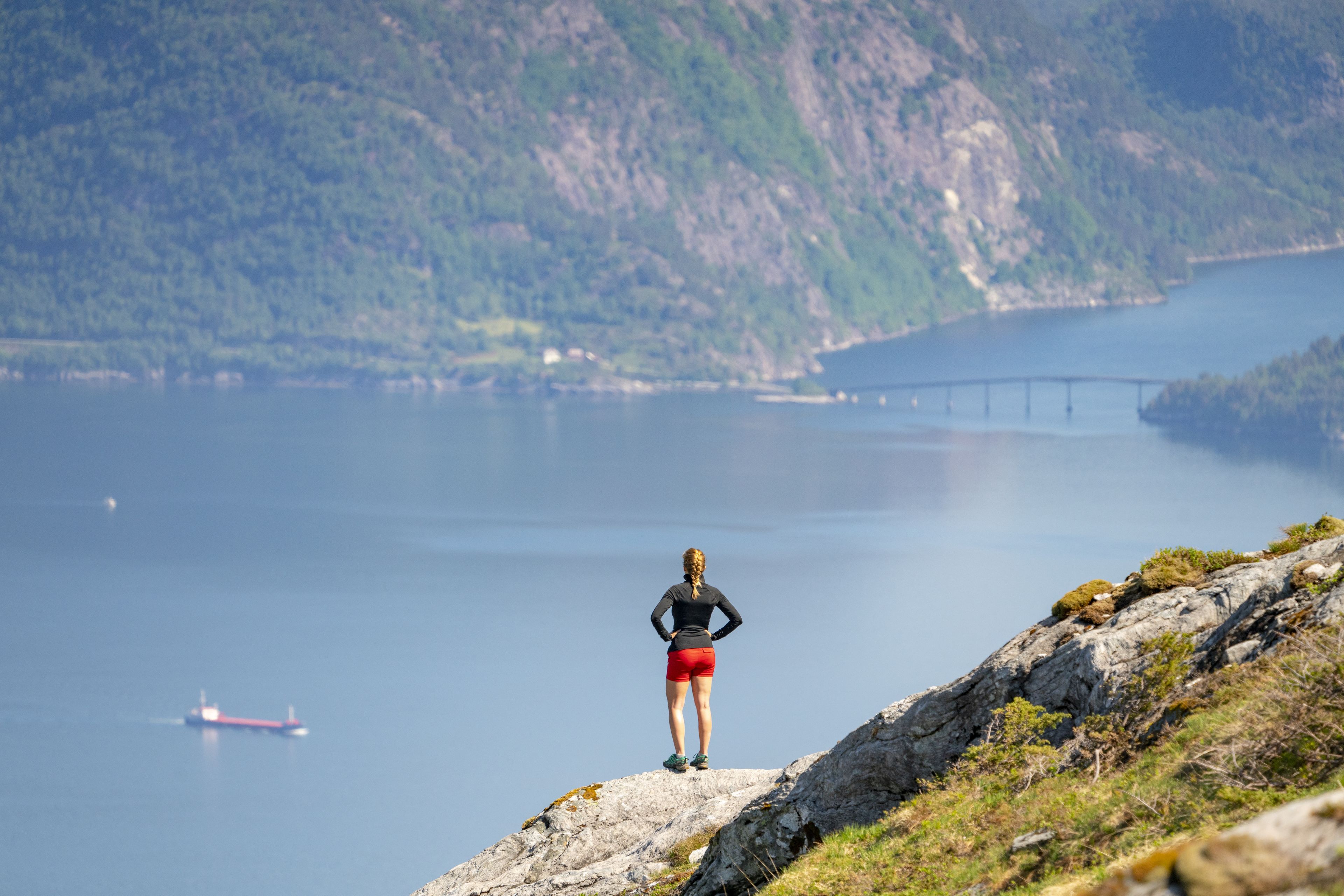A person standing on a rocky outcrop overlooking a calm body of water with a bridge in the distance.
