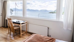 A room with a wooden desk, chair, and a bed with a brown blanket. A large window offers a view of a lake and mountains. - Lavik Fjord Hotell (Hoyanger)