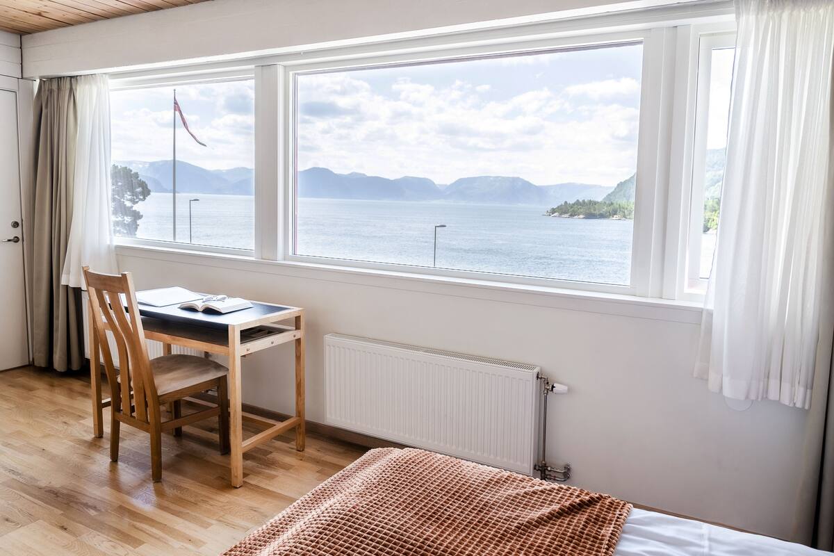 Una stanza con una scrivania in legno, una sedia e un letto con coperta marrone. Un'ampia finestra offre vista su un lago e montagne.