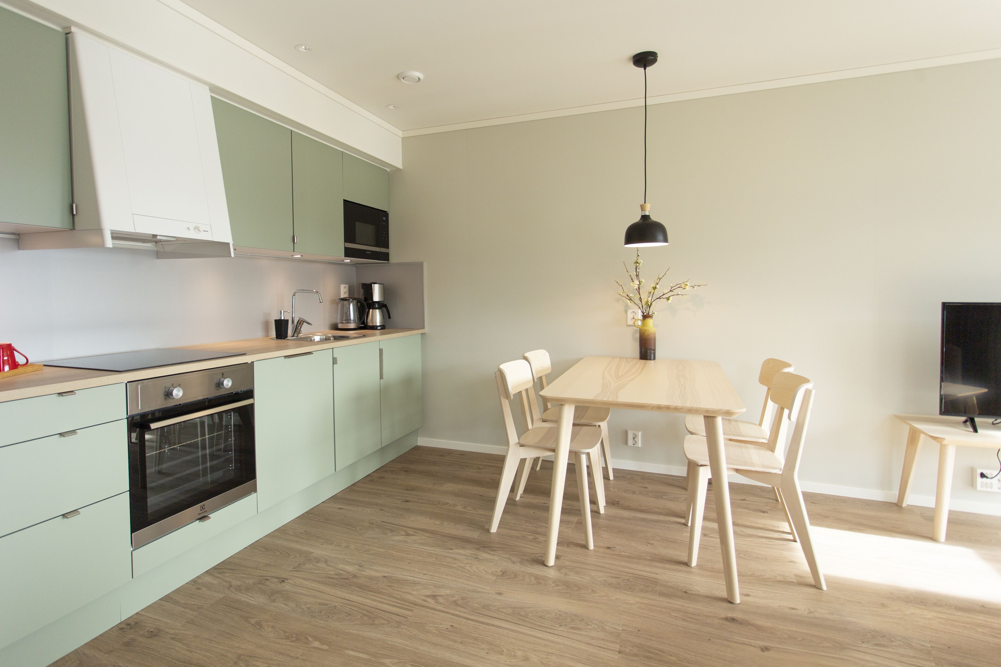 A modern kitchen with light green cabinets, a wooden dining table with four chairs, a coffee maker, and a TV on a stand.
