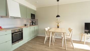 A modern kitchen with light green cabinets, a wooden dining table with four chairs, a coffee maker, and a TV on a stand.