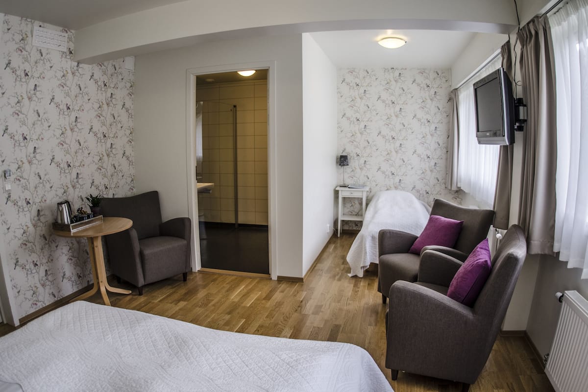 Camera d'albergo con un letto, due poltrone, un tavolino, una TV e un bagno visibile attraverso una porta aperta.