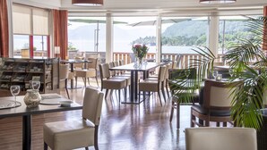 A restaurant with a view of a lake and mountains, featuring a long table with chairs, a bar area, and a bookshelf with magazines. - Lavik Fjord Hotell (Hoyanger)