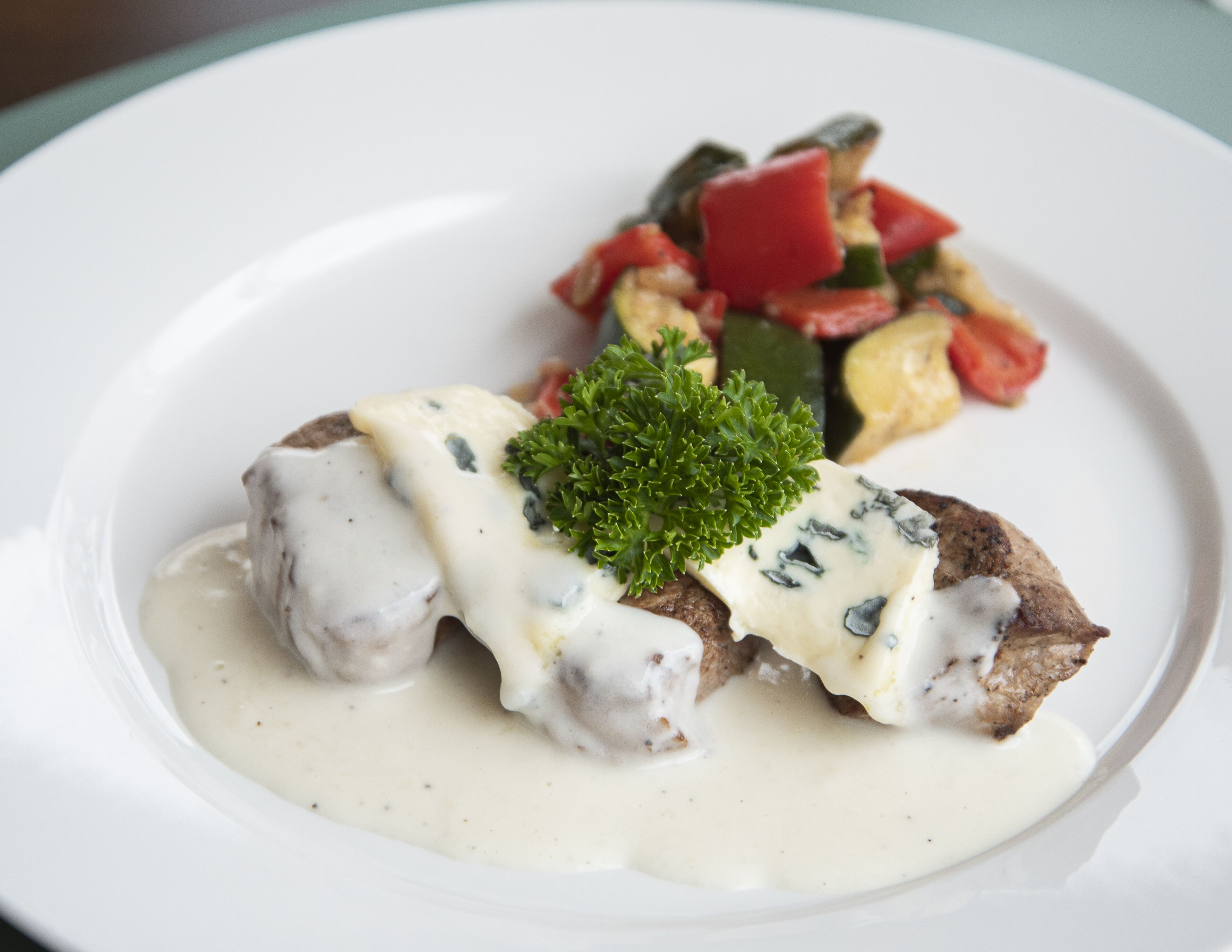 En tallrik med en kötträtt som är täckt med sås, garnerad med persilja och serverad med en blandning av grönsaker som tillbehör.