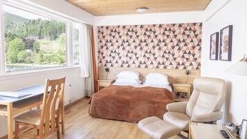 A bedroom with a wooden bed, a desk, a chair, and a view of a forest through a window.