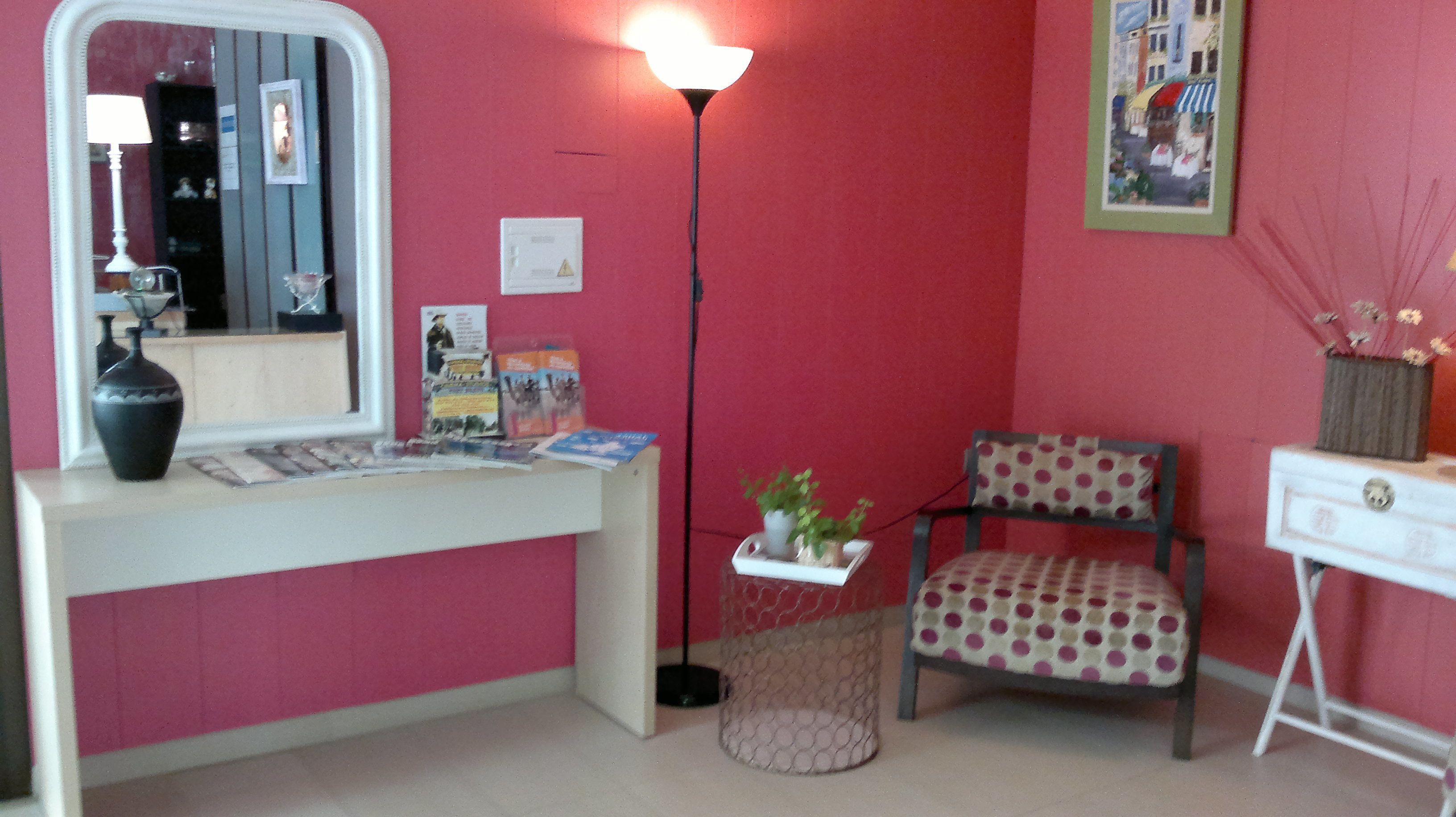 Een kamer met een roze muur, een wit bureau met spiegel, een lamp, een stoel en een klein tafeltje met een plant.
