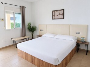 Habitación de hotel con una cama grande, un banco, una planta en maceta, una obra de arte colgada en la pared y una ventana con cortinas.
