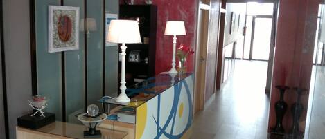 A hotel lobby with a reception desk, a bookshelf, a mirror, and a hallway leading to the exit.