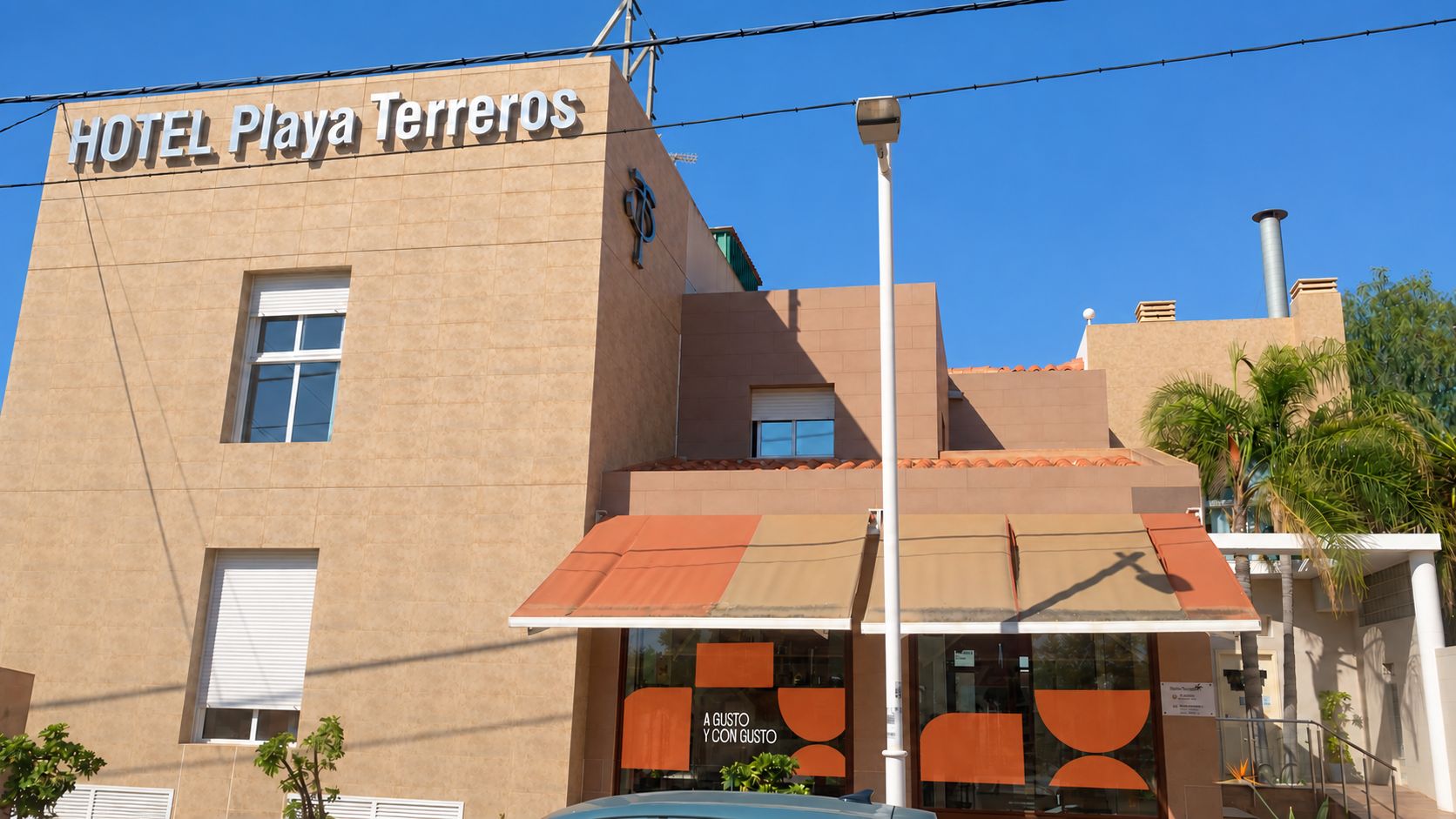 Un hotel llamado 'HOTEL Playa Terreros' con una fachada moderna, un acceso cubierto y un restaurante visible llamado 'A GUSTO YCCON GUSTO' en su interior.
