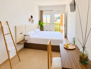 Un dormitorio con cama, una mesita de noche con un florero de vidrio, una silla y una ventana con vista a follaje.