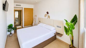 Una camera d'albergo con un letto grande, un comodino con telecomando, una TV a muro, un armadio in legno, un bagno con lavandino e specchio, e due piante in vaso.
