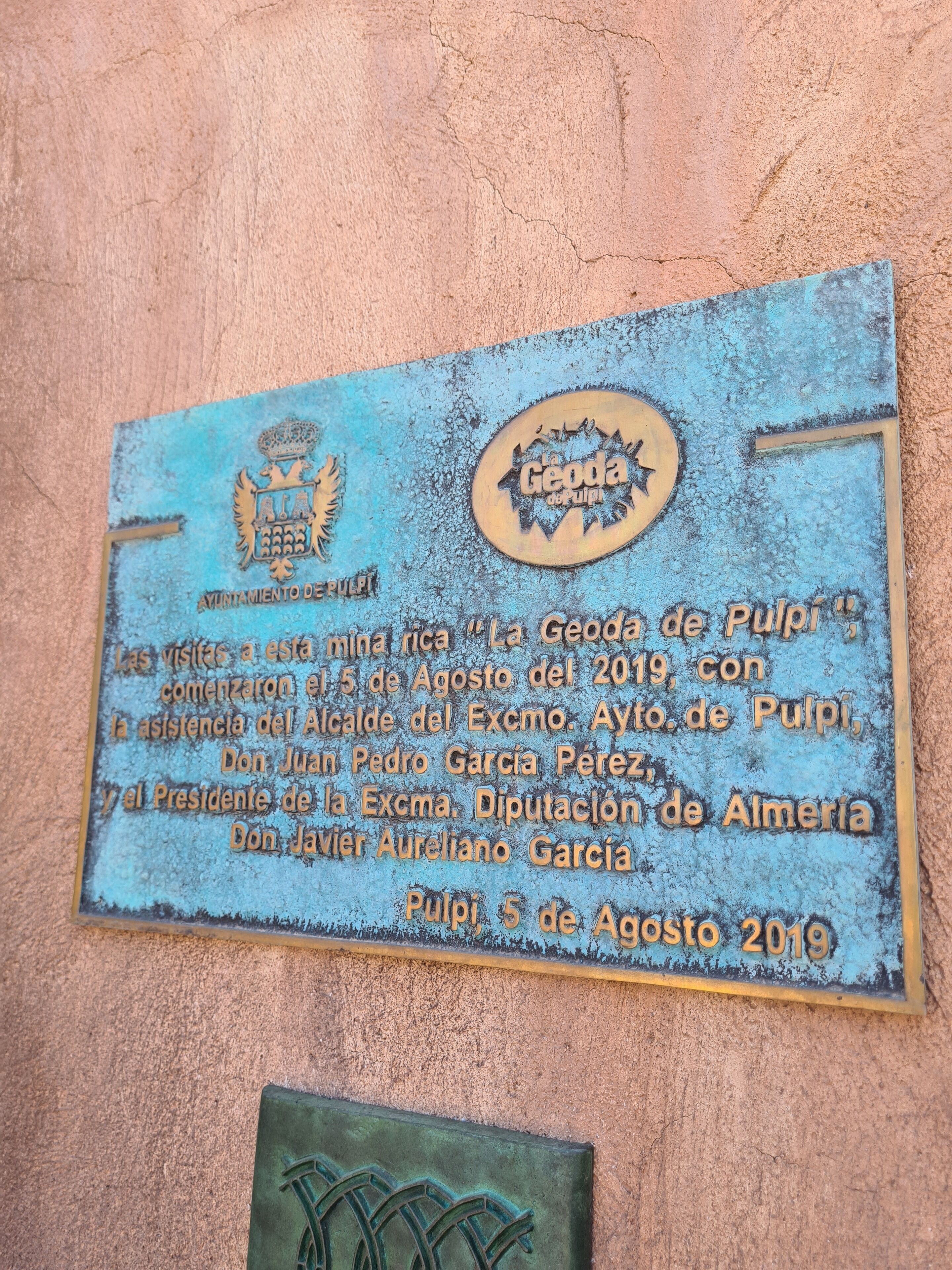 A weathered plaque with text and a crest, mounted on a textured wall.