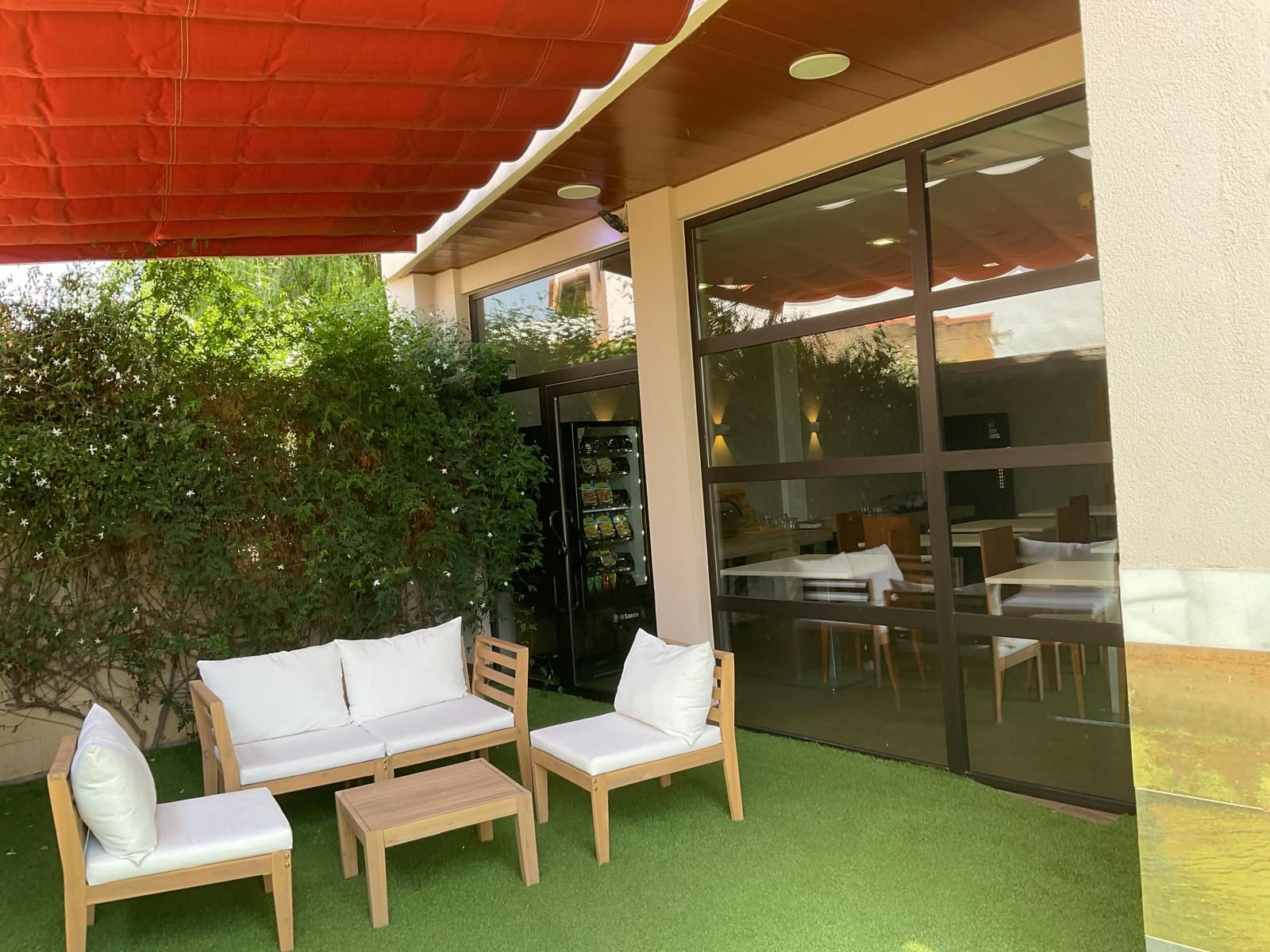 Een terras met een houten bank en stoelen, een bijzettafeltje en een glazen wand die het interieur weerspiegelt.