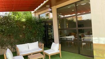 Een terras met een houten bank en stoelen, een bijzettafeltje en een glazen wand die het interieur weerspiegelt.