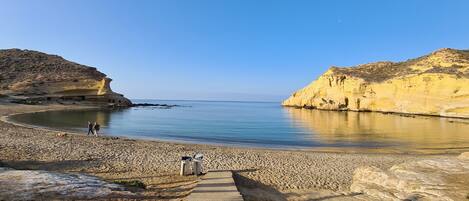 A coastal beach with a wooden walkway leading to the water, rocky cliffs, and a clear sky.