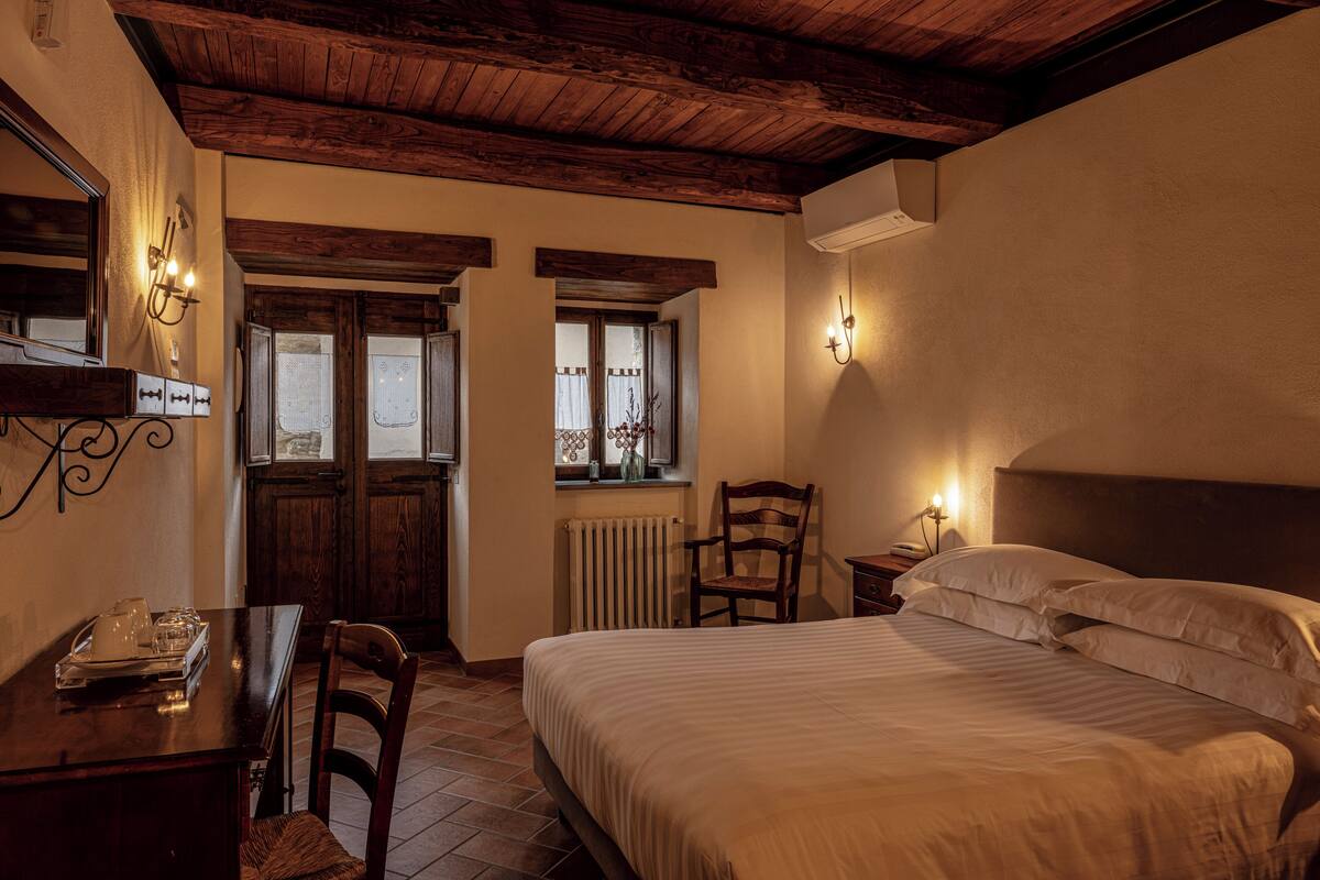 A bedroom with a wooden ceiling, a bed with white linens, a wooden chair, a small table with a tray, and a window with curtains.
