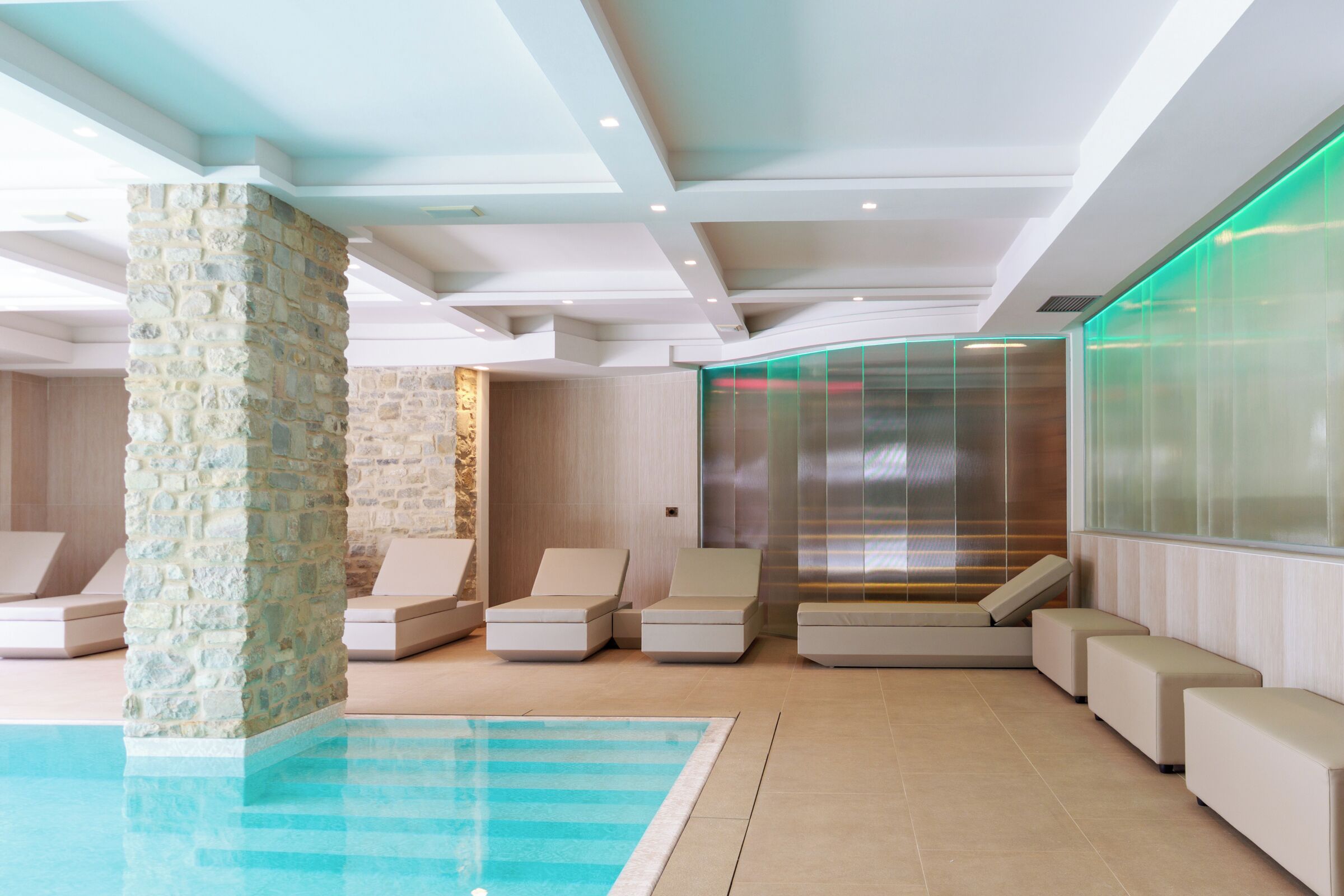 Un espace piscine intérieur moderne, doté de chaises longues, d’un pilier en pierre et d’un ascenseur en verre.