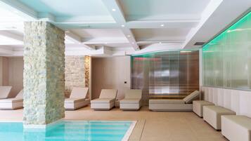 Un espace piscine intérieur moderne, doté de chaises longues, d’un pilier en pierre et d’un ascenseur en verre.