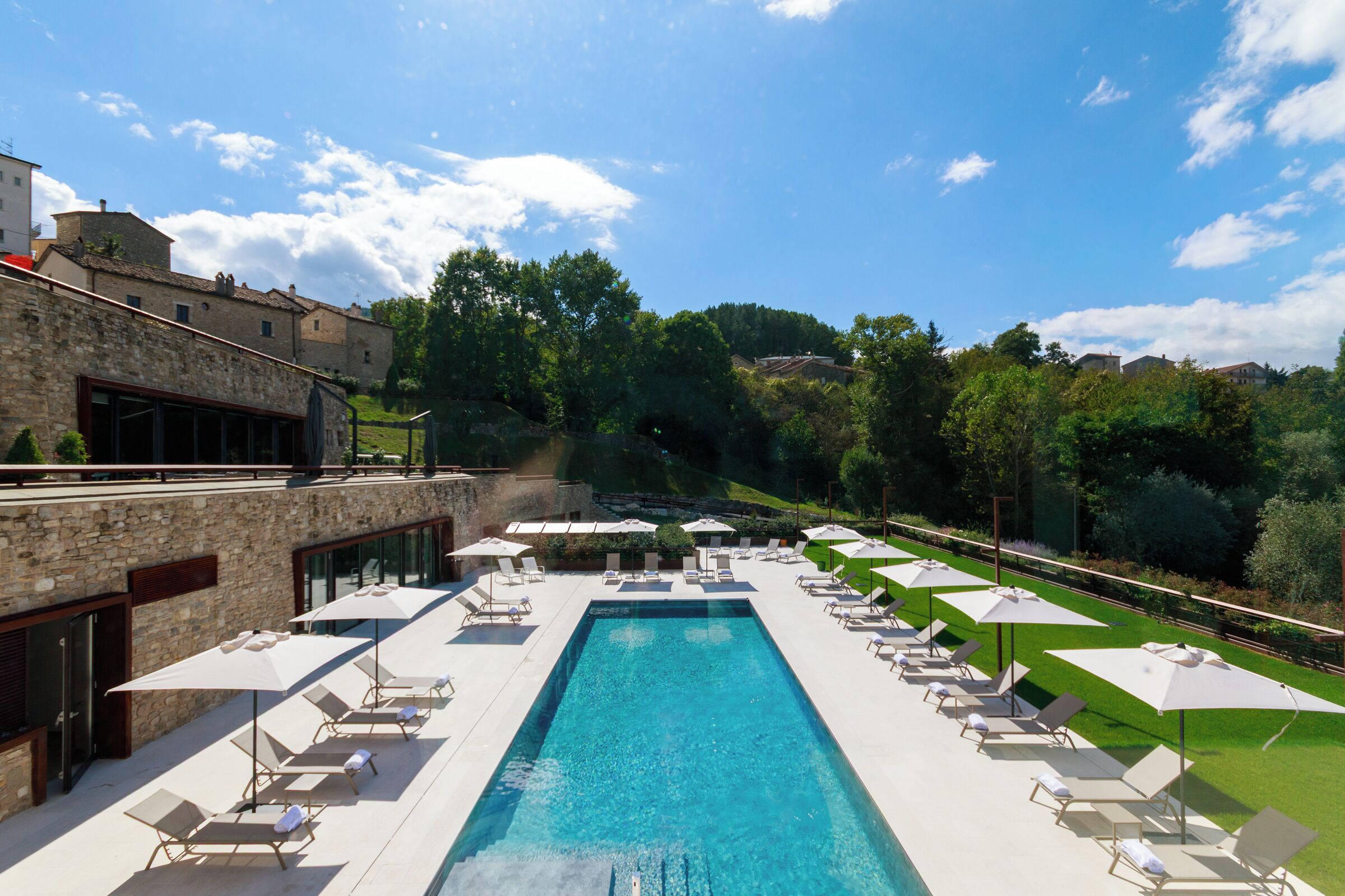 A swimming pool with lounge chairs and umbrellas, surrounded by greenery and buildings.