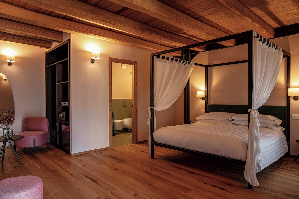 A four-poster bed with white linens, a wooden ceiling, and a small seating area with a pink chair.