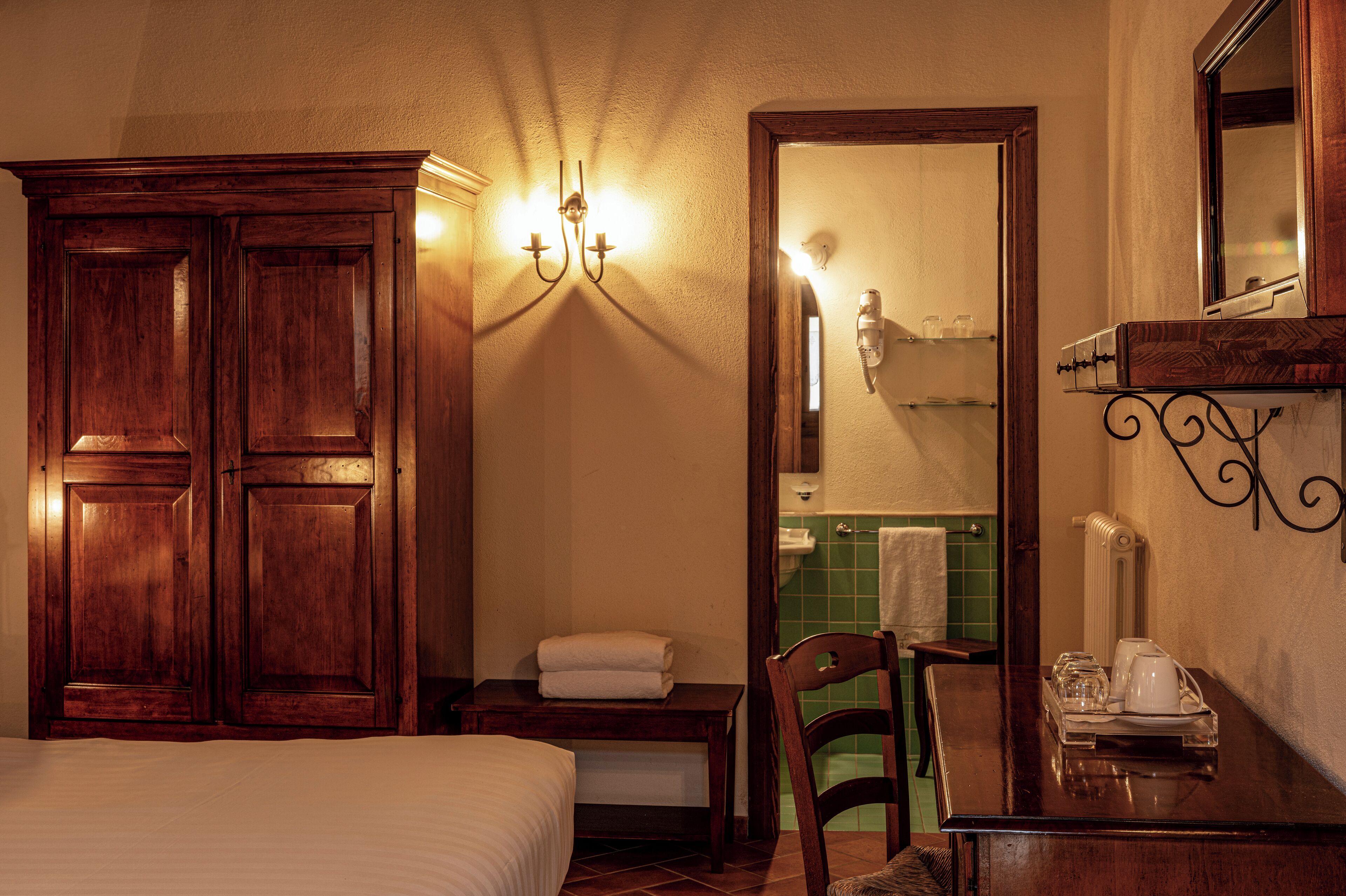 A hotel room with a wooden wardrobe, a bedside table with a lamp, a chair, and a small table with a tray of items.