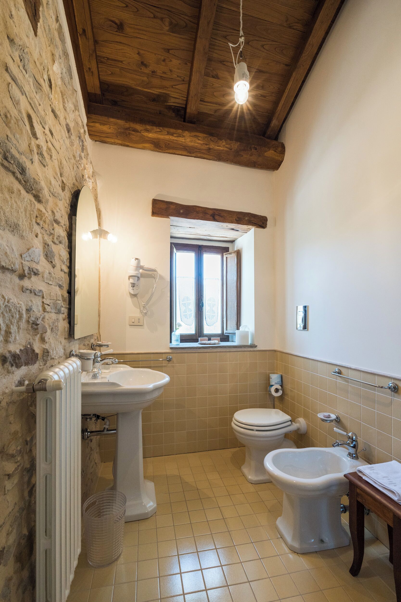A bathroom with a stone wall, wooden ceiling beams, a window, a sink, a toilet, and a bidet.