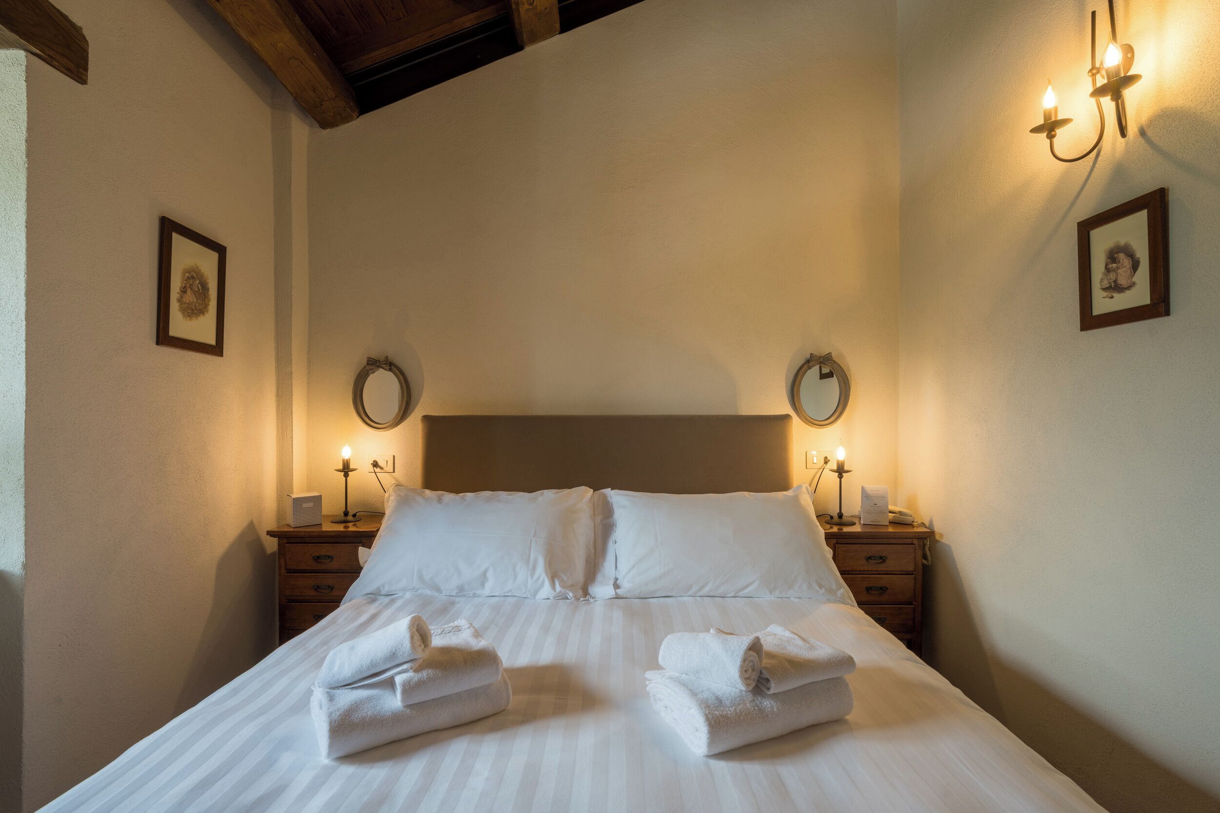A neatly made bed with white linens and towels, two bedside tables with lamps, and framed pictures on the walls.